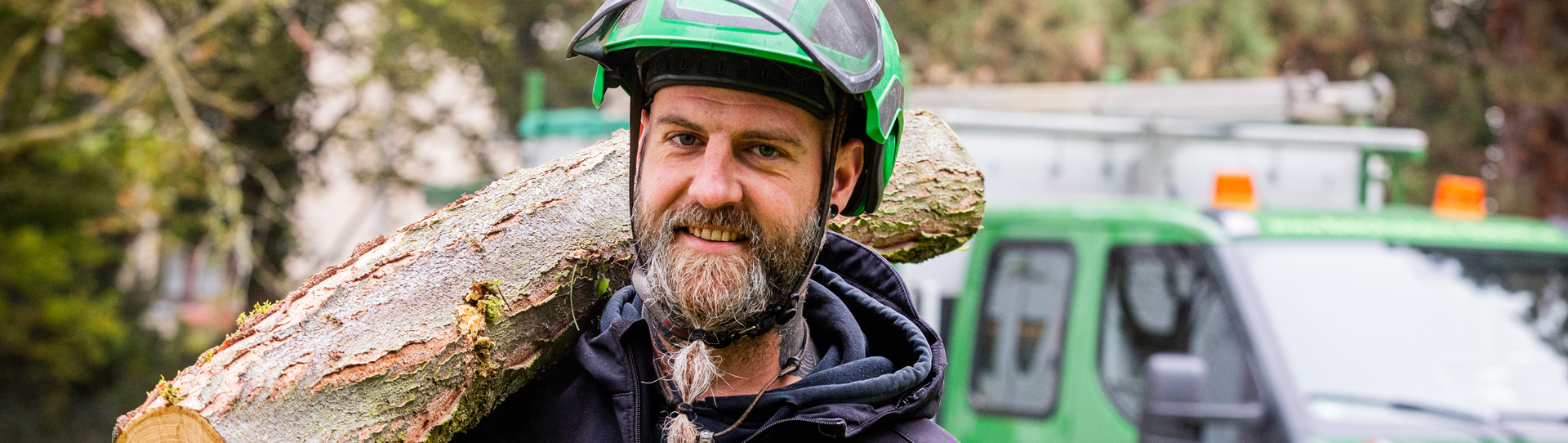 Baumpfleger trägt einen Baumstamm auf der Schulter. Im Hintergrund steht ein grünes Fahrzeug.